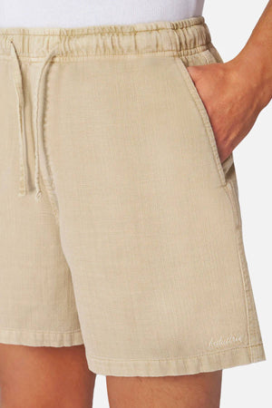 A person wears Industrie The Mallorca Short—men's relaxed-fit, light beige cotton shorts with subtle texture and small white embroidery near the hem—paired with a crisp white top, hand in the pocket.