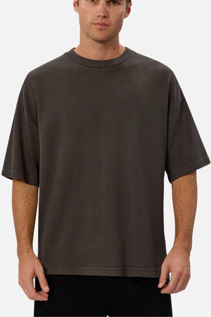 A man stands facing forward against a plain light background, wearing a relaxed fit, short-sleeve Industrie The Marlon Tee in dark gray paired with black pants.