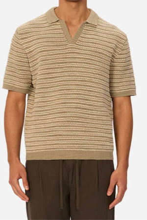 A man wearing the Industrie The Matera Polo—short-sleeved, beige and white striped, 100% cotton with a V-neck collar—and brown pants stands against a plain background.