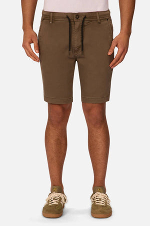 A person wearing Industrie The New Drifter Cuba Short—brown men's shorts with a black drawstring—paired with a white tucked-in shirt and olive green sneakers with cream laces, standing against a plain white background.