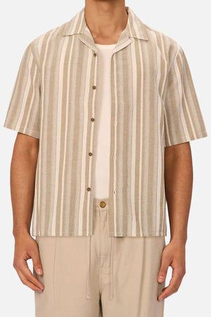 A man wears the Industrie The Textured Livorno Short Sleeve Shirt—beige and white vertical stripes, relaxed fit, short sleeves, camp collar—over a white T-shirt and light beige pants. Only his torso and arms are visible.