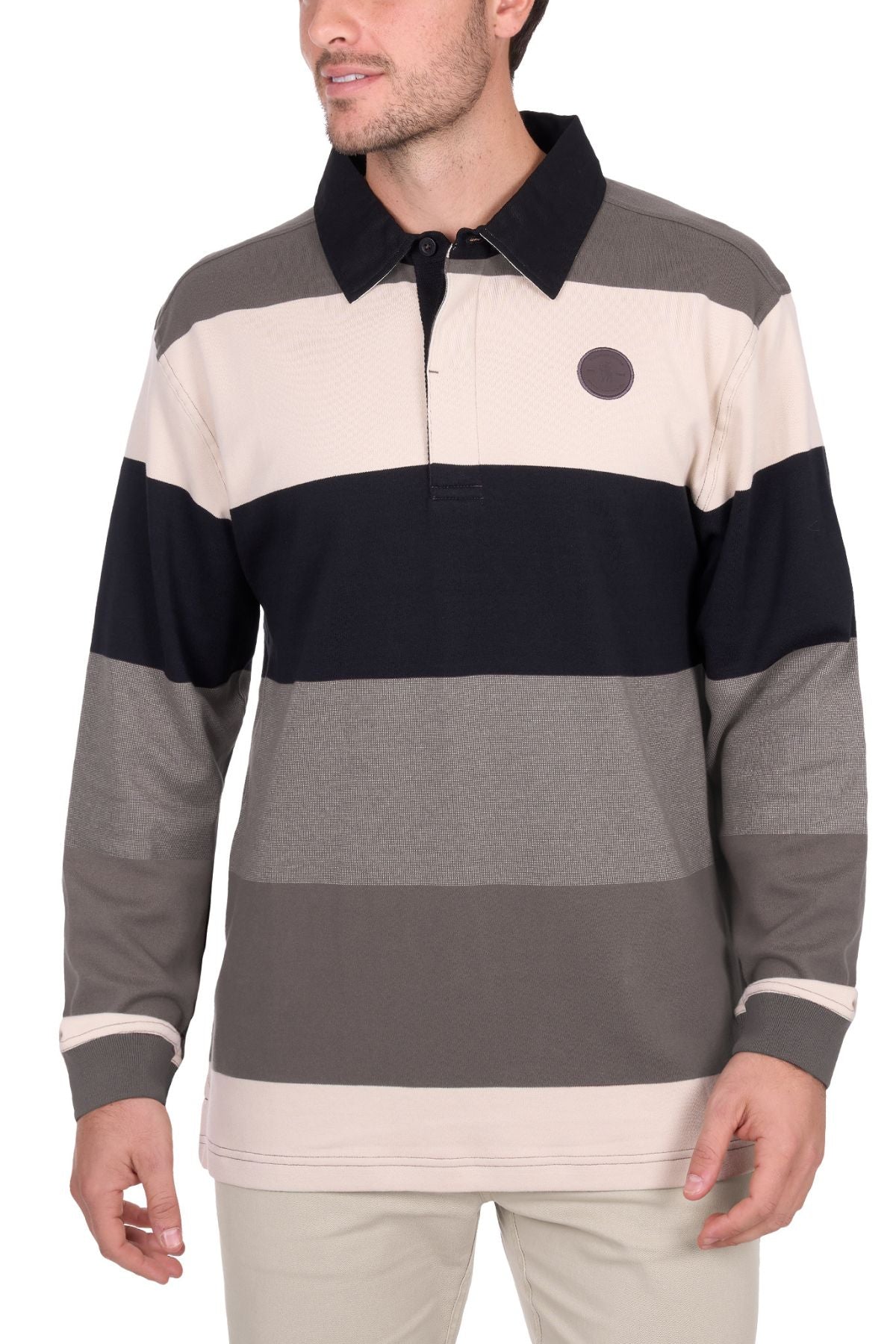 A man wears the Thomas Cook Brandon Rugby, a long-sleeve shirt with wide beige, black, and gray stripes, a collar, small circular chest patch, and light-colored pants.