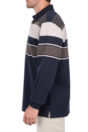 A man stands sideways with his left arm relaxed, wearing the Thomas Cook Bryan Stripe Rugby—a long-sleeve, collared shirt with horizontal stripes in navy blue, grey, beige, and brown.