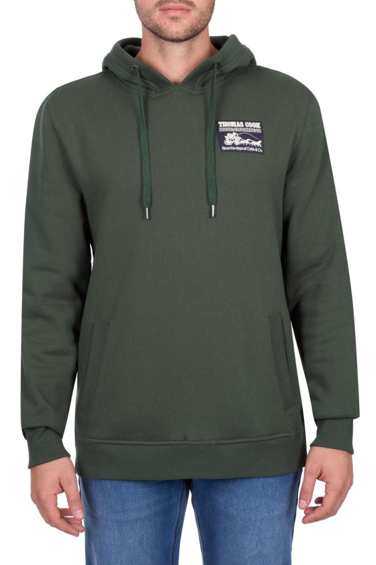 A man wears the Thomas Cook Heritage C N C Hoodie, made from heavyweight brushed fleece with a "Thomas Cook" chest patch, and blue jeans, standing against a plain white background.