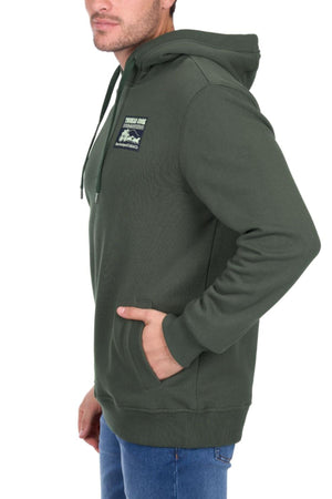 A man wearing the Thomas Cook Heritage C N C Hoodie in dark green with a chest patch and blue jeans stands sideways, one hand in his pocket.