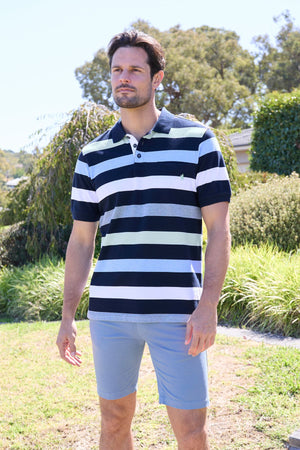 A man stands outdoors among greenery and trees on a sunny day, wearing the Thomas Cook Charles Polo—a 100% cotton pique multi-colour stripe polo in navy, white, and light blue—paired with matching light blue shorts.