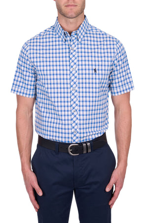 A man wears a Thomas Cook Chris Tailored Short Sleeve Shirt, blue and white checkered, tucked into dark navy pants with a black belt, standing against a white background.