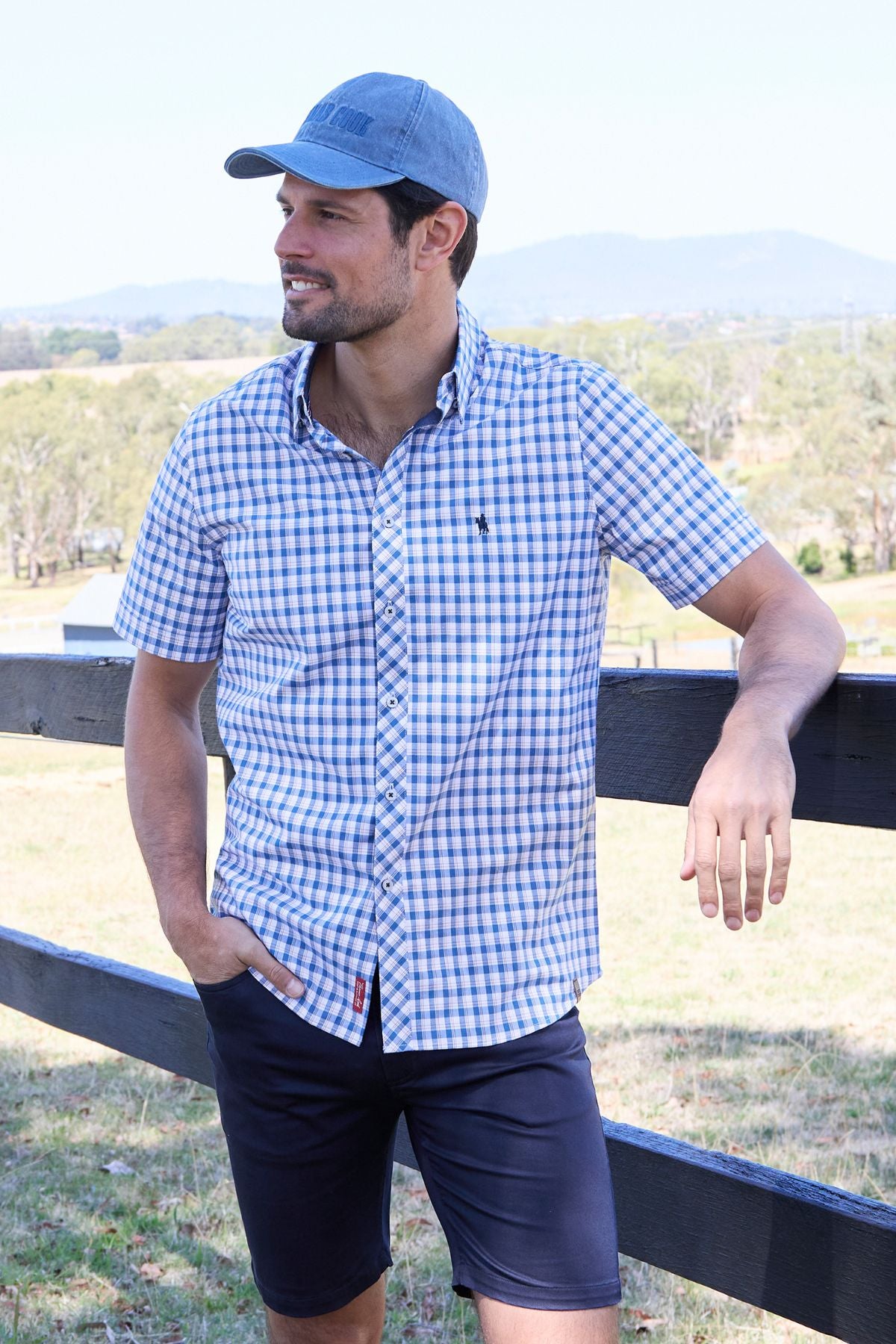 A man in the Thomas Cook Chris Tailored Short Sleeve Shirt, navy shorts, and a blue cap stands outdoors by a wooden fence, smiling with a scenic landscape in the background.