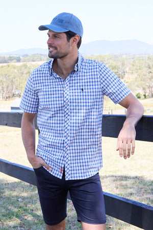 A man in the Thomas Cook Chris Tailored Short Sleeve Shirt, navy shorts, and a blue cap stands outdoors by a wooden fence, smiling with a scenic landscape in the background.