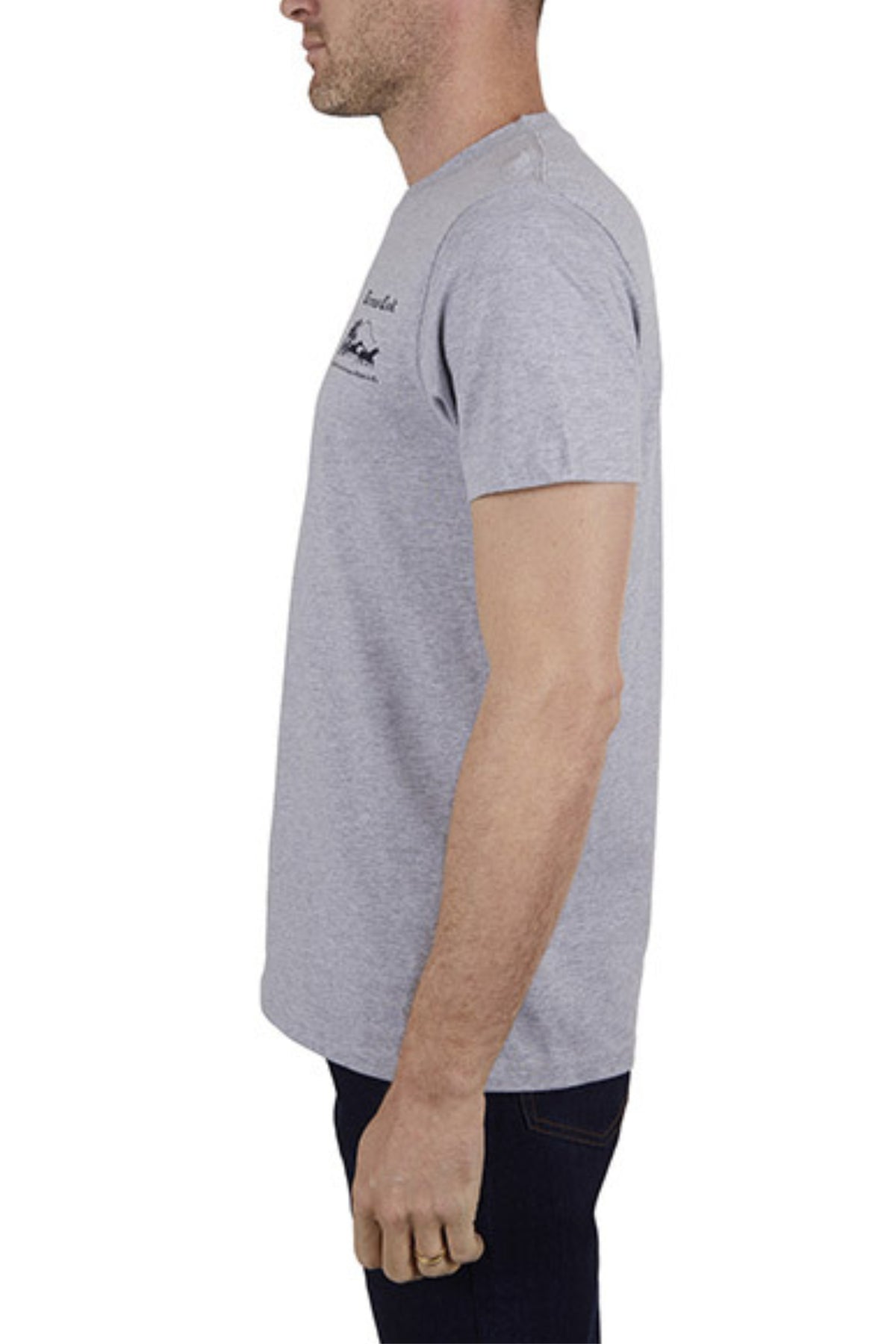 A man in profile stands against a plain white background, wearing the Thomas Cook Coach Tee in light grey and dark jeans, arms relaxed at his sides.