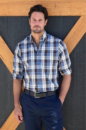 A man with dark hair and a beard stands before a black wall with a wooden X pattern, wearing the Thomas Cook Daniel Shirt with rolled-up sleeves and navy blue pants.