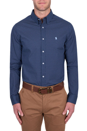 A man wears the Thomas Cook David Tailored Shirt, a navy blue geometric print, long-sleeve button-down, paired with khaki pants and a brown belt, standing against a plain white background.
