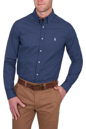 A man wears the Thomas Cook David Tailored Shirt, featuring a dark blue geometric print and small chest logo, tucked into light brown pants with a brown belt.