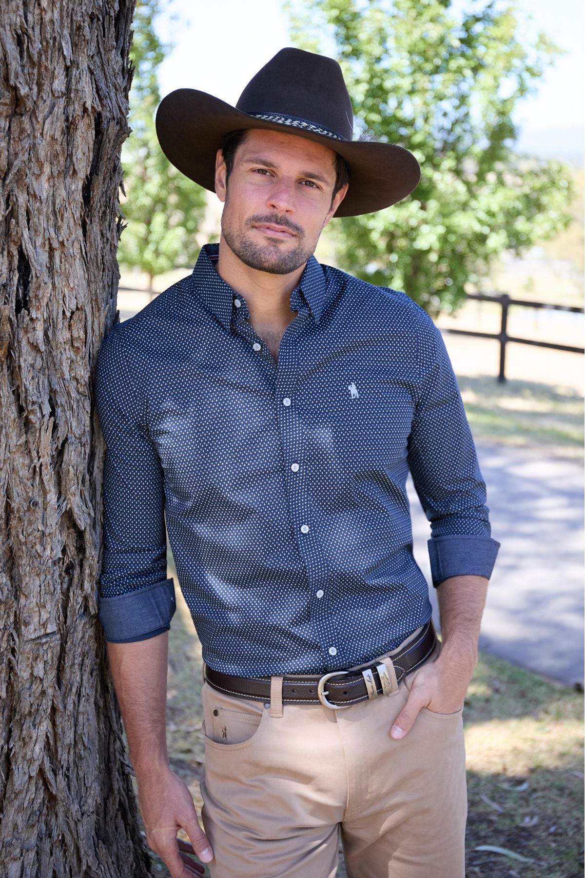 A man in a Thomas Cook David Tailored Shirt, tan pants, and a wide-brimmed brown hat leans against a tree near a wooden fence and green trees on a sunny day.