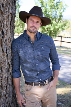 A man in a Thomas Cook David Tailored Shirt, tan pants, and a wide-brimmed brown hat leans against a tree near a wooden fence and green trees on a sunny day.