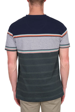 A man stands facing away from the camera, wearing a Thomas Cook Dion Tee—an all-cotton jersey tee featuring navy, gray, and green multi-colour stripes—and light-colored pants.