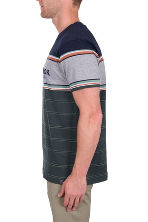 A man shown from the side wears the Thomas Cook Dion Tee, a 100% cotton jersey short-sleeved tee with navy, gray, and dark green color-blocked stripes. Only his upper body is visible; he also wears beige pants.