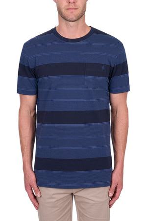 A man stands facing forward against a white background wearing the Thomas Cook Edward Tee—navy blue with multi-colour stripes, chest pocket, and beige pants. Made from 100% cotton jersey for comfort.