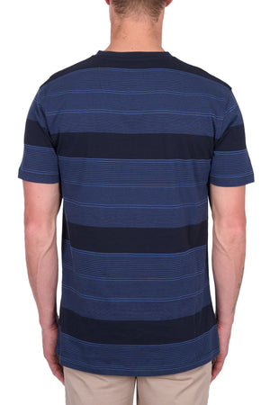 A person is seen from behind wearing the Thomas Cook Edward Tee, a short-sleeve navy blue t-shirt with light blue stripes in 100% cotton jersey, paired with beige pants against a plain white background.