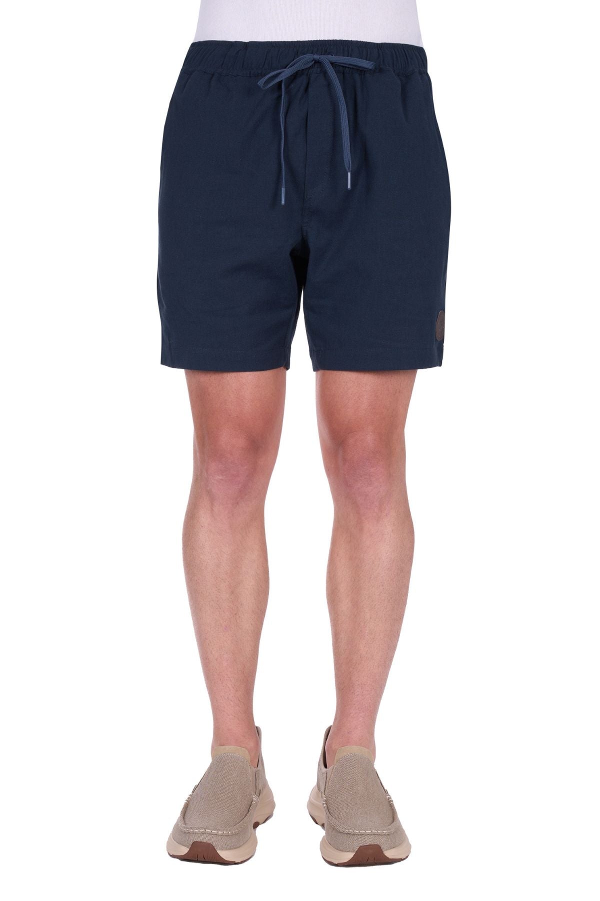 A person is seen from the waist down wearing Thomas Cook Felix Linen Shorts in navy blue with beige slip-on shoes, standing against a white background.
