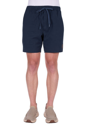 A person is seen from the waist down wearing Thomas Cook Felix Linen Shorts in navy blue with beige slip-on shoes, standing against a white background.