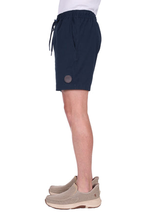 A man in side profile wears the Thomas Cook Felix Linen Short in navy, featuring a round patch on the left leg and beige slip-on shoes, photographed against a white background.