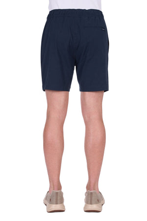 A person is shown from the back against a plain white background, wearing navy blue Thomas Cook Felix Linen Shorts, a white shirt, and beige slip-on shoes.