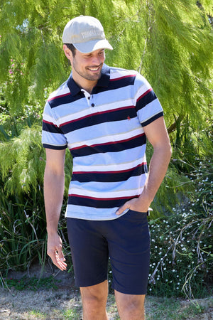 A man wearing a Thomas Cook Gavin Polo in multicolour stripes, a beige cap, and navy shorts stands outdoors smiling, with green trees and bushes in the background.