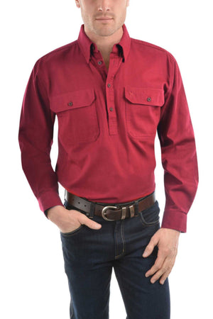 A man wears a long-sleeved, red Thomas Cook Heavy Drill Half Placket Shirt with two chest pockets, dark blue jeans, and a brown belt, standing with one hand in his pocket against a white background.