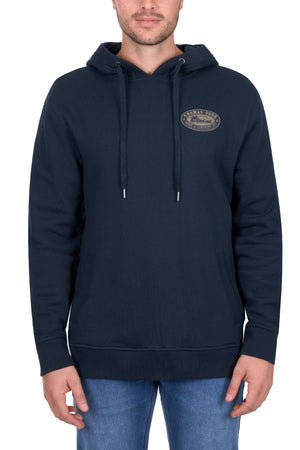A man faces forward, wearing the Thomas Cook Heritage Oval Hoodie in navy with a small left-chest emblem, drawstrings, and front pocket, paired with light blue jeans. The background is plain white.