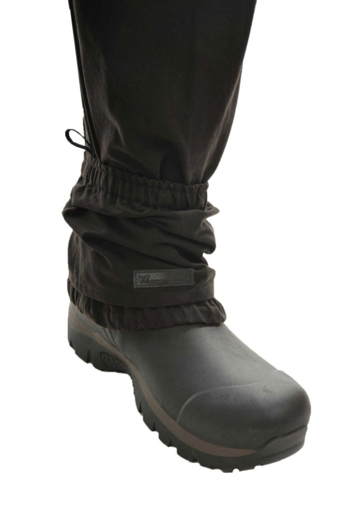 A close-up of a person’s lower leg wearing Thomas Cook High Country Bushman Short Oilskin Gaiters over black pants and sturdy hiking boots for outdoor protection, isolated on a white background.