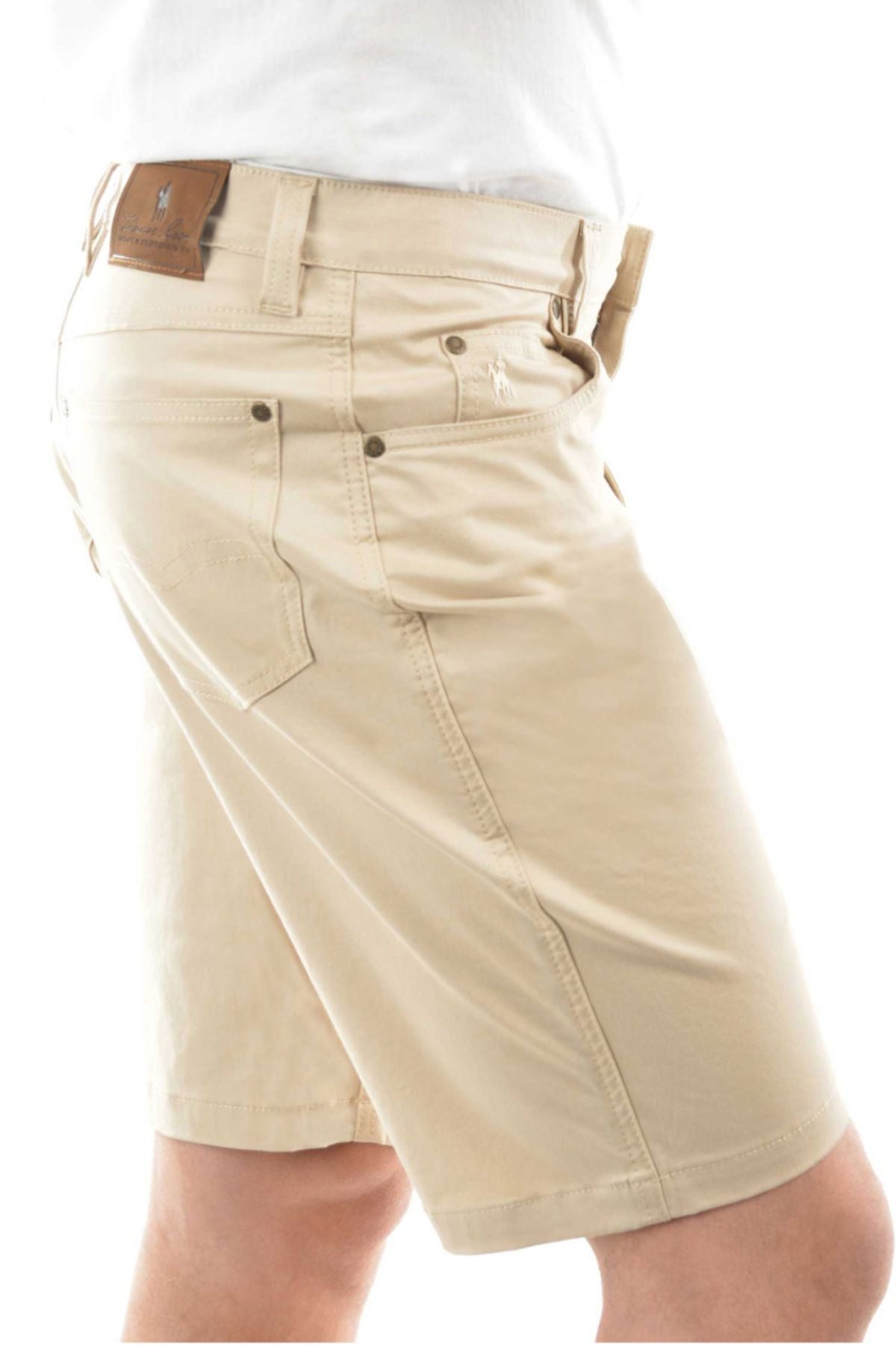A person wearing Thomas Cook Jake Comfort Waist Shorts in light beige stretch fabric with side and back pockets, belt loops, a brown leather logo patch on the waistband, paired with a white shirt. Side view photo.