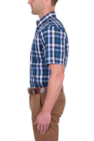 A man is shown in profile wearing the Thomas Cook John Short Sleeve Shirt, featuring a blue and white check pattern, tucked into tan pants with a brown belt. The background is plain white.