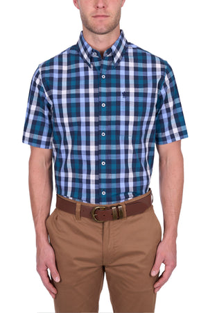 A man wears the Thomas Cook John Short Sleeve Shirt in blue, teal, and white cotton checks, buttoned up and tucked into tan pants with a brown belt, standing against a white background.