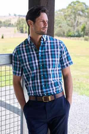 A man in a Thomas Cook John Short Sleeve Shirt and navy pants stands outdoors, leaning against a fence and post, gazing thoughtfully at the lush greenery and trees in the background.