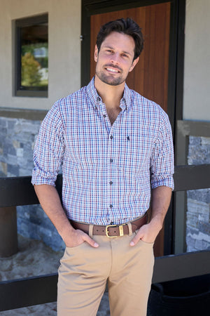 A man with brown hair and a beard smiles outdoors, wearing the Thomas Cook Joseph Shirt in 100% easy-care cotton. His plaid button-down has rolled sleeves, paired with khaki pants, as he stands before a stone and wood building.