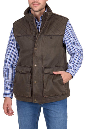 A man wears the Thomas Cook Marcus Vest in brown over a blue and white plaid shirt and blue jeans, with one hand in his vest pocket, shown from the thighs up against a white background.