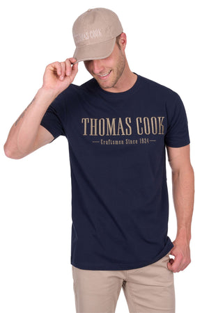 A man in a beige cap and navy blue Thomas Cook Nathan Tee, made from 100% cotton jersey with chest embroidery, smiles while touching his cap brim and wears beige pants.
