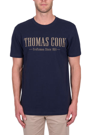 A man wears the Thomas Cook Nathan Tee in navy, made from 100% cotton jersey with "THOMAS COOK Craftsmen Since 1924" embroidered on the chest, styled with light beige pants against a plain white background.
