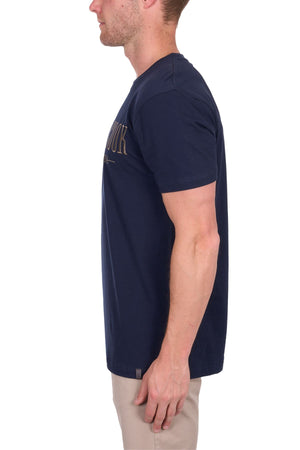 A man in profile wears a navy Thomas Cook Nathan Tee with chest embroidery and beige pants, standing against a plain white background.