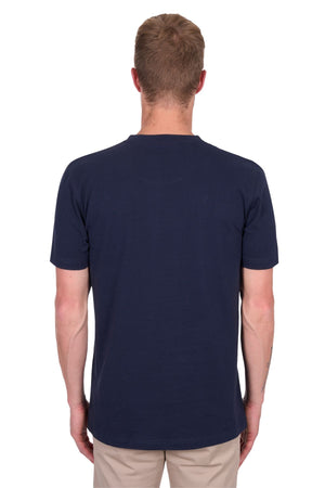 A person with short light brown hair stands with their back to the camera, wearing a navy Thomas Cook Nathan Tee and beige pants against a white background.