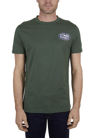 A man stands against a plain white background, wearing the Thomas Cook Oval Tee in dark green—a classic fit, 100% cotton t-shirt featuring a small blue and white oval logo on the left chest.