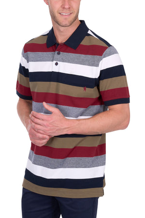 A man stands with his hands clasped, wearing the Thomas Cook Sean Polo— a cotton pique shirt featuring wide stripes in maroon, olive green, white, grey, and navy blue—paired with dark pants.