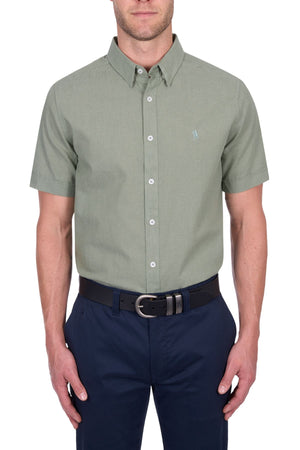 A man wears the Thomas Cook TC Linen Short Sleeve Shirt in light green, tucked into dark blue pants with a black belt, standing against a white background—an effortless summer essential.