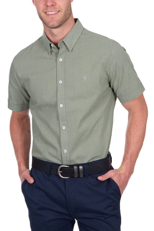 A man wears the Thomas Cook TC Linen Short Sleeve Shirt in light green with dark blue pants and a black belt, hands in pockets and smiling—a relaxed summer essential, shown cropped from the shoulders down.