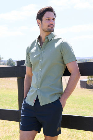 A man stands outdoors by a black wooden fence in the Thomas Cook TC Linen Short Sleeve Shirt, light green and perfect for sunny days—a true summer essential.