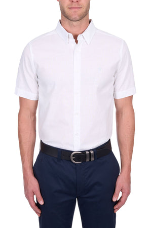 A man stands with arms relaxed at his sides against a white background, wearing the Thomas Cook TC Linen Short Sleeve Shirt in white, paired with navy blue pants and a black belt.