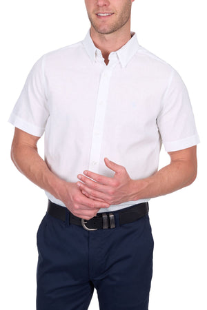 A man wears the Thomas Cook TC Linen Short Sleeve Shirt in white, tucked into navy blue pants with a black belt, standing against a white background. His face is partially out of frame.