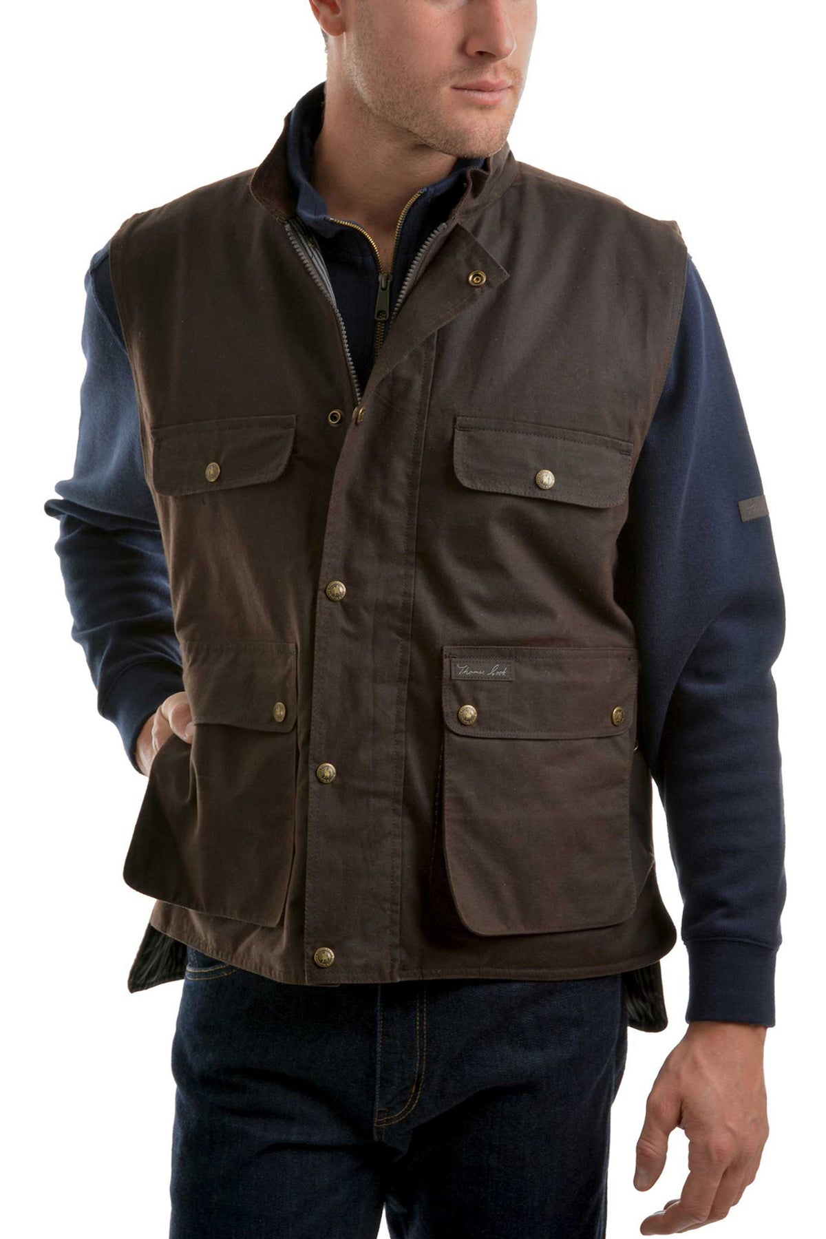 A man wears the Thomas Cook High Country Oilskin Vest, a dark brown waxed cotton vest with multiple pockets, brass buttons, and a zip front, over a navy long-sleeve shirt and dark jeans while facing slightly to the side.