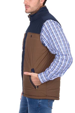 A man wears the Thomas Cook Toby Revs Vest, a brown and navy sleeveless vest, over a blue and white plaid shirt with blue jeans, standing with his left hand in the vest pocket against a white background.